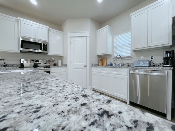A kitchen with white cabinets, a granite countertop, stainless steel appliances, and a window with blinds.