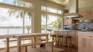 A kitchen with wooden cabinets, a large window overlooking a body of water, and a bar area with stools. - Oceanfront Paradise: Breathtaking 3bd/2ba with Panoramic Views of Hilo Bay (Hilo)