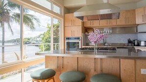 A modern kitchen with wooden cabinets, a stainless steel range, and a large window overlooking a body of water. - Oceanfront Paradise: Breathtaking 3bd/2ba with Panoramic Views of Hilo Bay (Hilo)