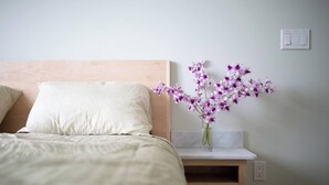 A neatly made bed with a white pillow and a wooden headboard. A vase with purple flowers is placed on a small wooden shelf beside the bed. - Oceanfront Paradise: Breathtaking 3bd/2ba with Panoramic Views of Hilo Bay (Hilo)