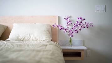 Ein ordentlich bezogenes Bett mit einem weißen Kopfkissen und einem hölzernen Kopfteil. Auf einem kleinen Holzregal neben dem Bett steht eine Vase mit violetten Blumen.