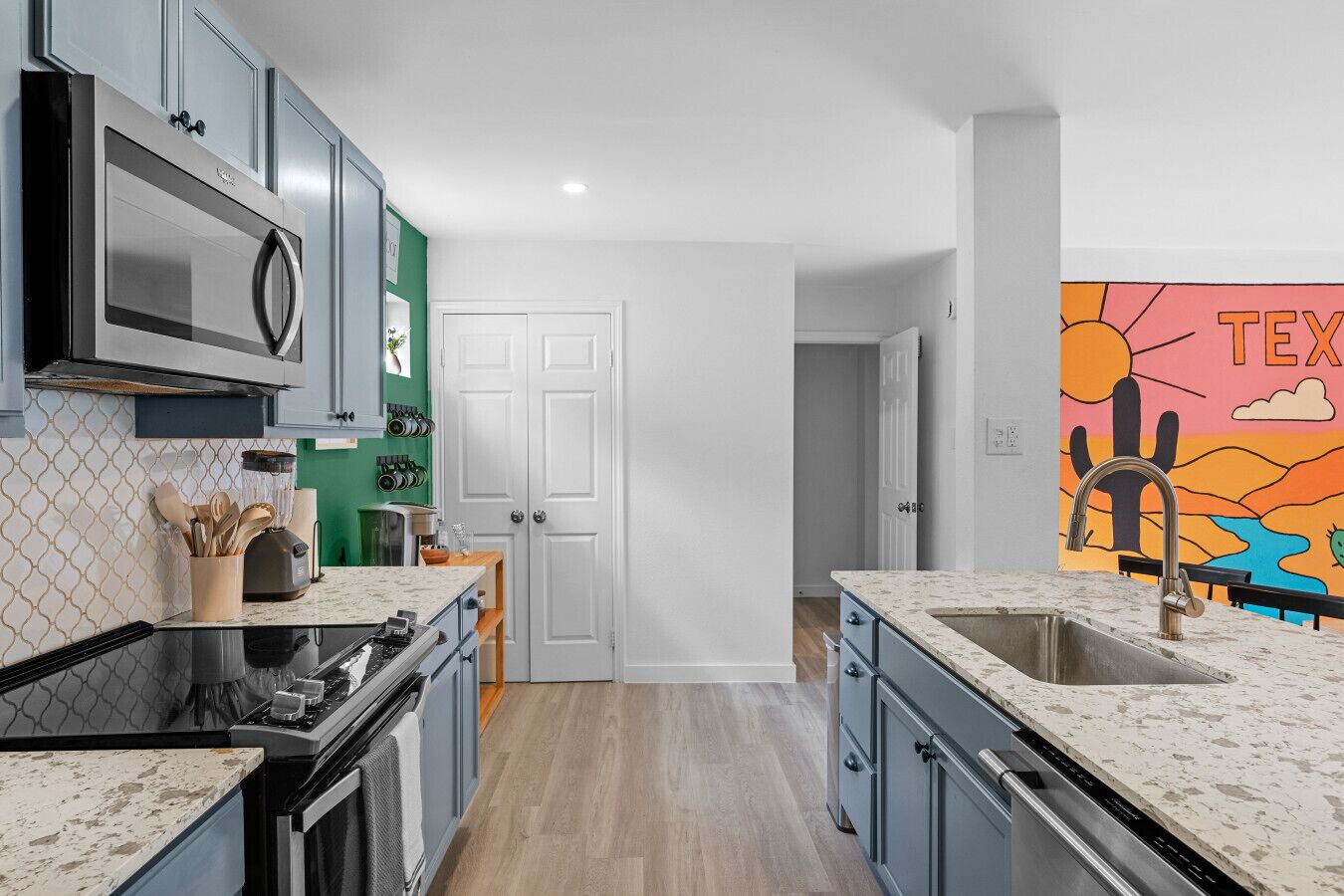 Une cuisine moderne équipée d’un four à micro-ondes, d’une cuisinière et d’un évier. Une porte donne accès à une autre pièce et un mur est décoré d’une fresque colorée.