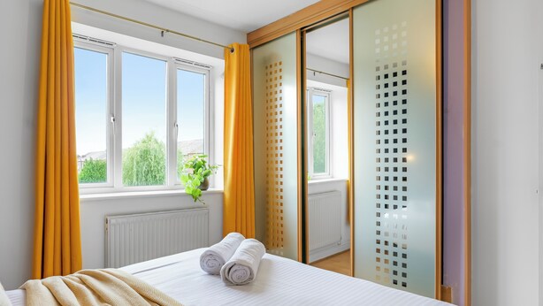 A hotel room with a bed, yellow curtains, a window with a view, and a mirror with a decorative panel.