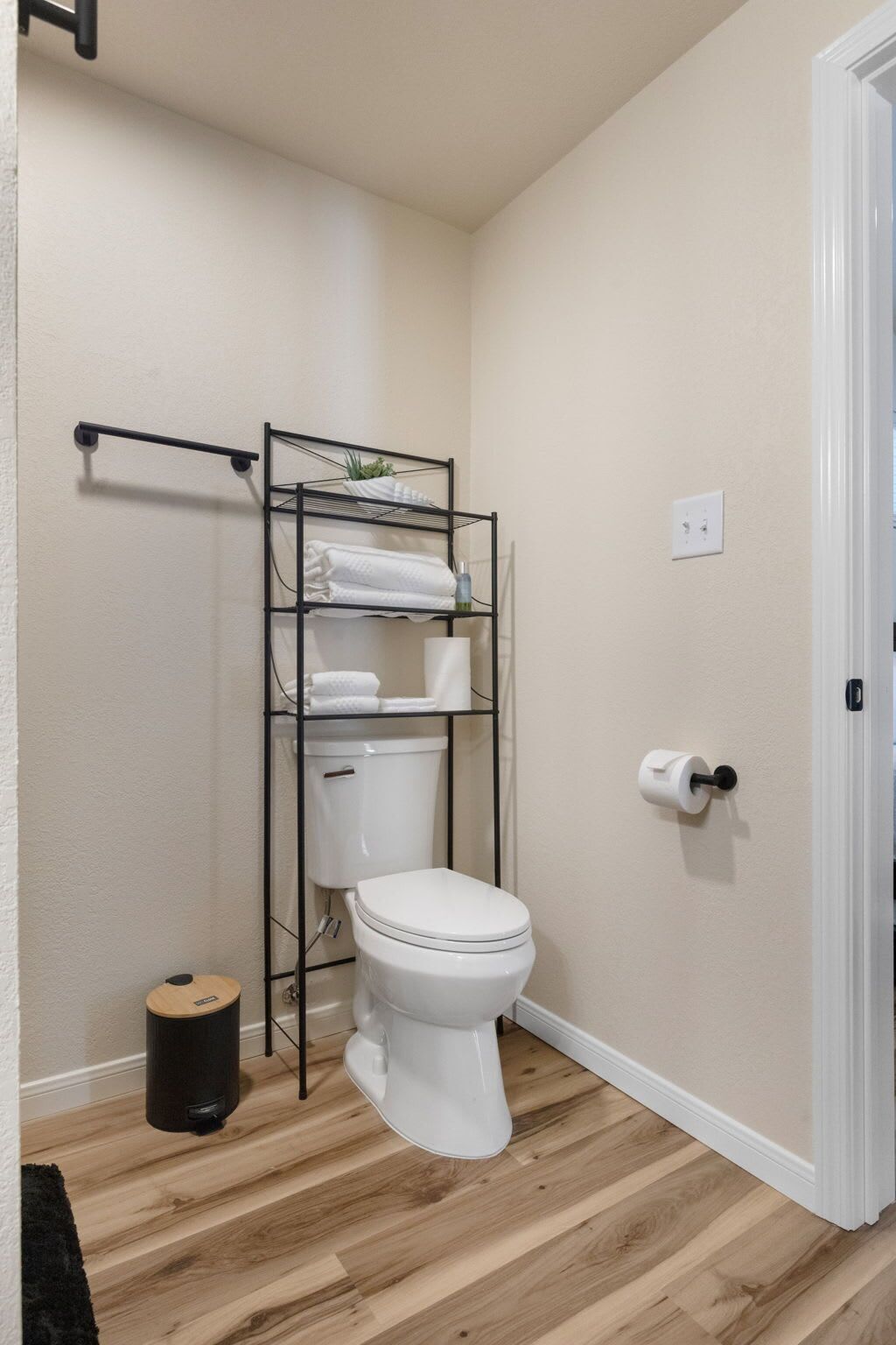 A small bathroom with a white toilet, a black metal shelf unit with towels, a black trash bin, and a white door.