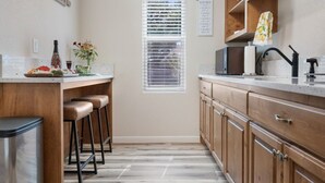 A kitchen with wooden cabinets, a stainless steel trash can, a window with blinds, and a countertop with a microwave. - Kleines Hauschen - Cozy Hideaway 6 min to Downtown (Fredericksburg)
