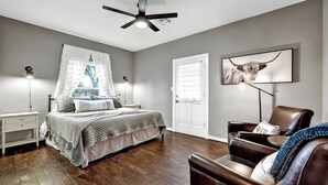 A bedroom with a bed, bedside tables, a ceiling fan, a chair, and a wall-mounted TV. - The Country Haus: Quaint Home 1 Mile From Downtown (Fredericksburg)