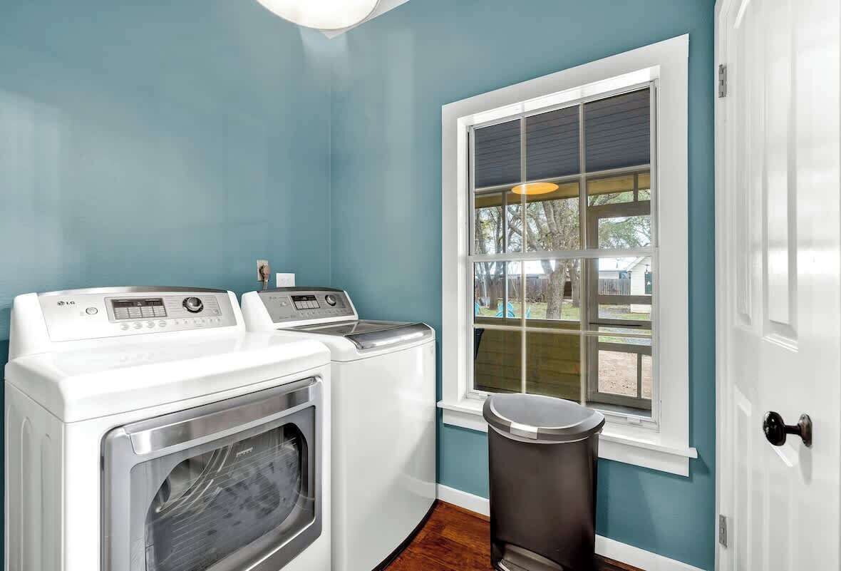A laundry room with a front-loading washer and dryer, a window with a view of the outdoors, and a trash can.