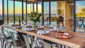 Een eetruimte met een lange tafel gedekt voor zes personen, grote ramen met uitzicht op het landschap, en een vaas met bloemen op tafel.