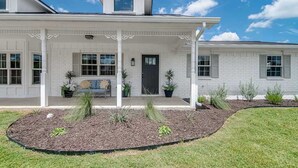 A white two-story house with a covered porch, a black door, and a small garden area with plants and mulch. - 108 Wisteria Dr (Fredericksburg)