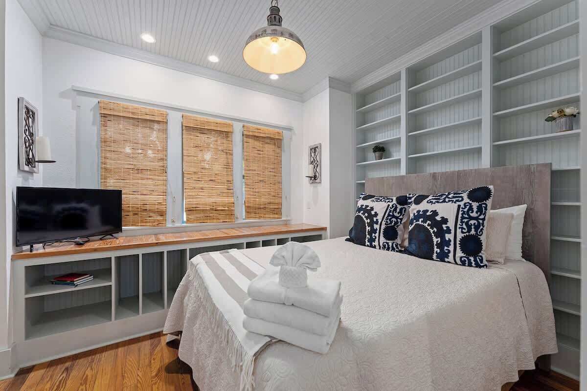 Un dormitorio con cama, televisión, estante con libros y ventana con persianas.