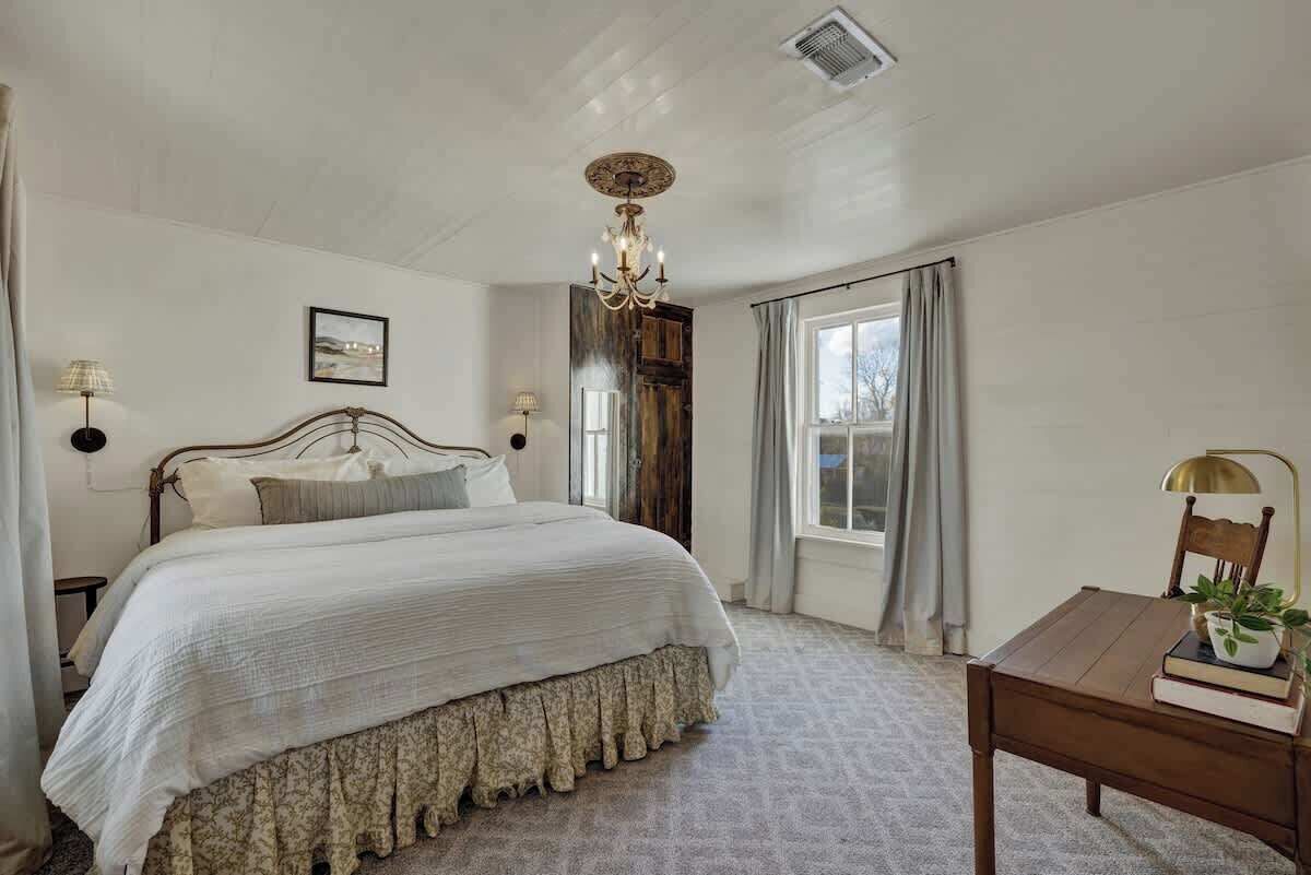 A bedroom with a large bed, a wooden headboard, a chandelier, a window with curtains, a bedside table with a lamp, and a small table with a plant and books.
