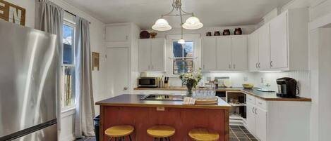 Una cocina con alacenas blancas, un refrigerador de acero inoxidable, una isla de madera con taburetes y una lámpara colgante.