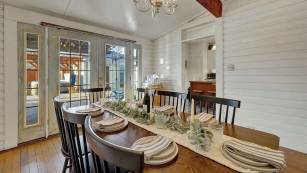 Un comedor con una larga mesa de madera dispuesta para seis personas, rodeada de sillas negras. Un candelabro cuelga del techo, y las paredes de la habitación están revestidas de paneles blancos.