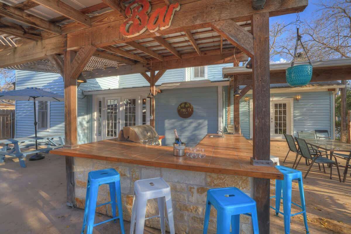 An outdoor bar area with a stone countertop, blue stools, and a wooden pergola overhead.