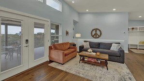 A spacious living room with a grey couch, a brown leather sofa, and a wooden coffee table. There are large windows and a view of the outdoors. - Esperanza Ranch W/hot-tub, Fire Pit, Gorgeous View (Fredericksburg)