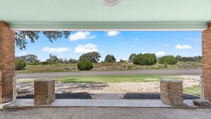 A covered outdoor area with a gravel floor, two brick pillars, and a view of a road and trees in the distance. - Hill Country Relaxstation Getaway (Fredericksburg)