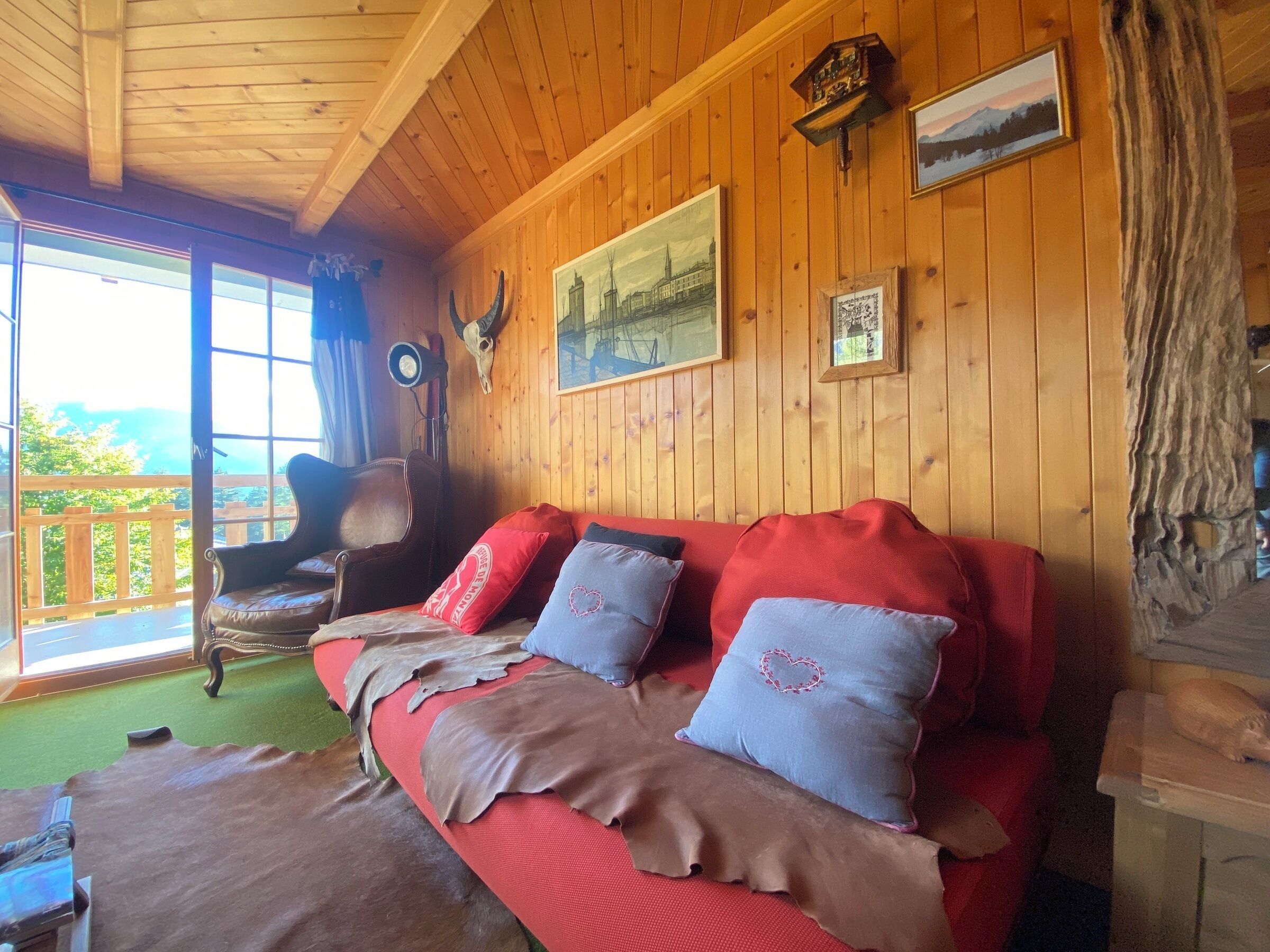Interior acogedor de una cabaña con techo de madera, un sofá rojo con cojines de cuero, un cráneo de venado montado y un cuadro con un paisaje.