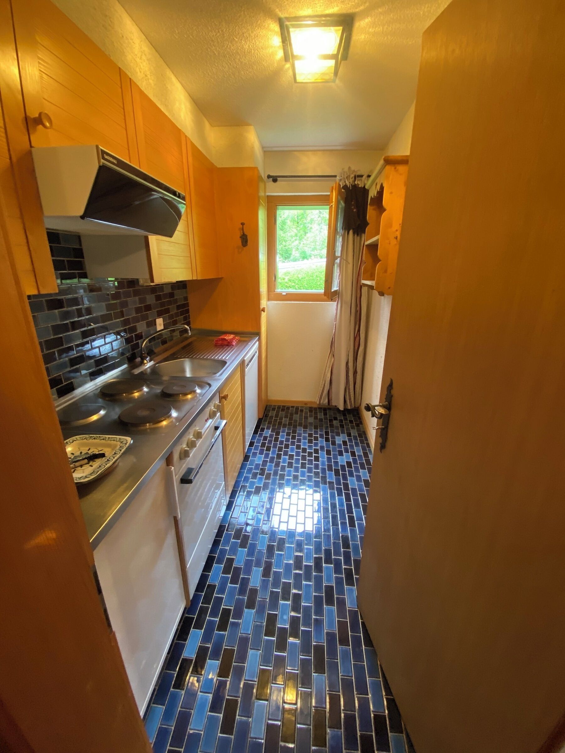 Une cuisine étroite avec un sol carrelé, des meubles en bois, une cuisinière en acier inoxydable et une fenêtre donnant sur un paysage verdoyant.