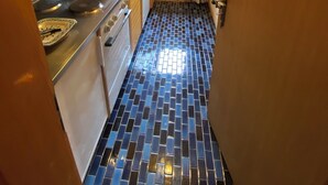 A narrow kitchen with a tiled floor, wooden cabinets, a stainless steel stove, and a window with a view of greenery. - The apartment is in a small 3-storey residence and is ideally located in a quiet area just above the (Gryon)