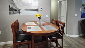 Un comedor con una mesa redonda de madera, cuatro sillas, un jarrón con flores amarillas y un cuadro de paisaje grande en la pared.