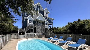 Une piscine avec des chaises longues, une terrasse en bois et une maison à deux étages avec un balcon en arrière-plan.