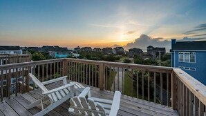 Une terrasse en bois avec des fauteuils Adirondack blancs, donnant sur un quartier résidentiel au coucher du soleil.