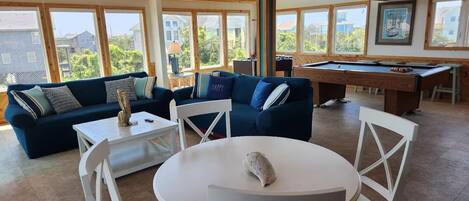 A spacious living area with a dark blue sofa set, a pool table, and a round table with chairs. Large windows provide ample natural light and views of the surrounding neighborhood.
