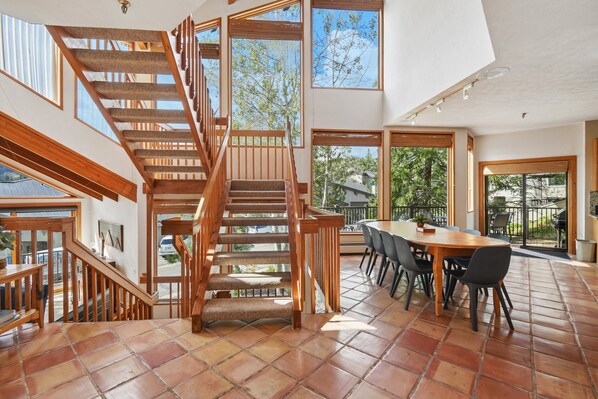 A spacious dining area with a high wooden staircase leading to an upper level, large windows offering views of the outdoors, and a tiled floor.