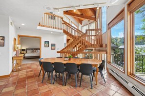 Un vaste espace dédié à la salle à manger, avec une table et des chaises en bois, un escalier doté de rampes en bois et de grandes fenêtres offrant une vue sur l’extérieur.