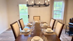 Una sala da pranzo con un tavolo in legno apparecchiato per sei persone, circondato da sedie imbottite. Un lampadario pende dal soffitto, e la stanza ha ampie finestre con tende.