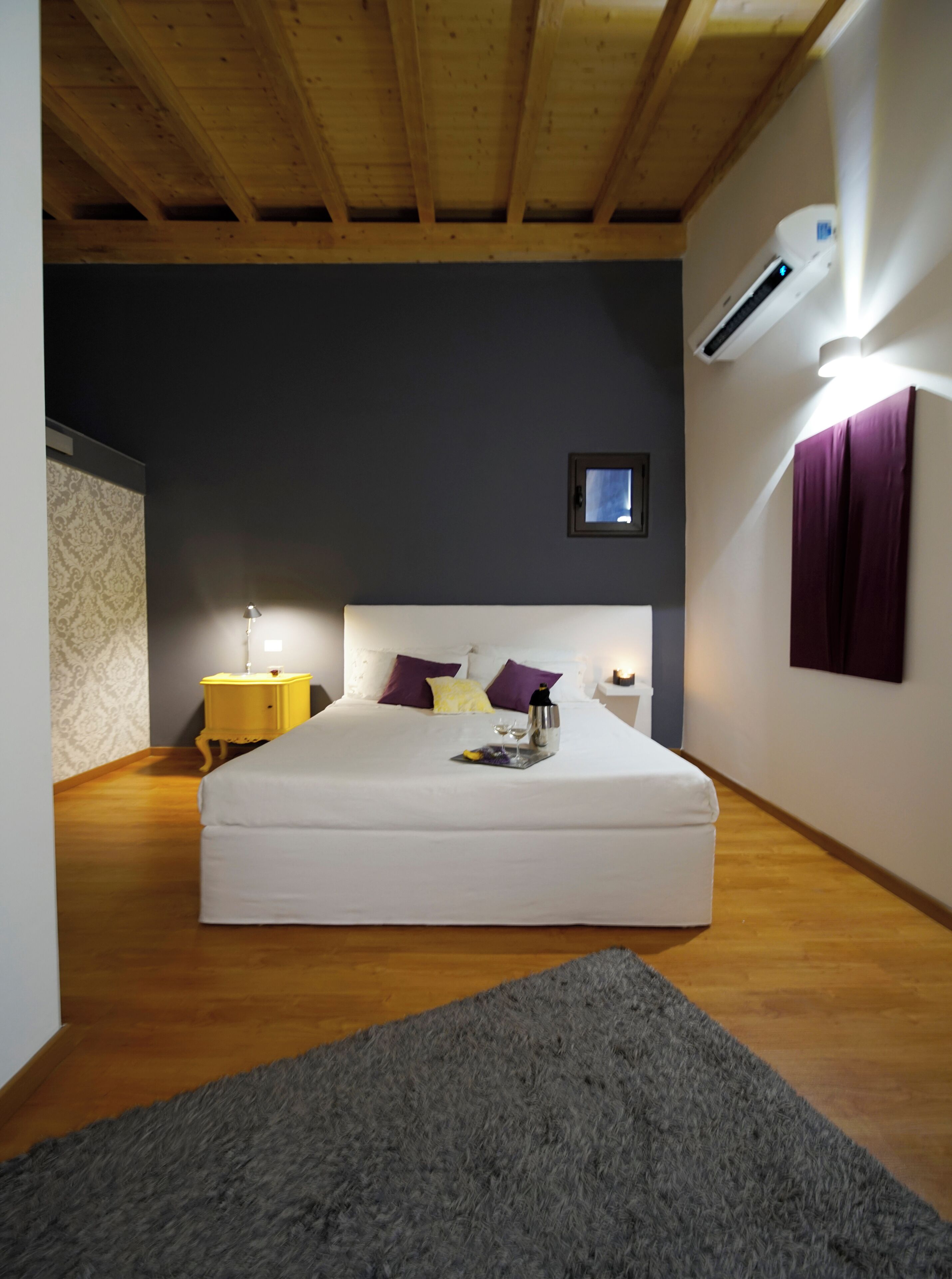 Une chambre avec un plafond en bois, un lit avec une literie blanche, une table de chevet, une lampe, une petite fenêtre et un appareil de climatisation.