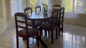 A dining area with a round table and six chairs, large windows with curtains, and a tiled floor.