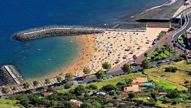 An aerial view of a beach with a rocky breakwater, a sandy shore, and a road leading to a parking area. - Charming retreat in Machico - 5 min Beach and B Center (Machico)