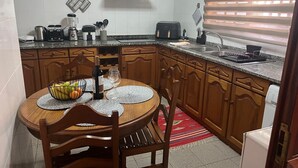 A kitchen with a round wooden table, chairs, a fruit bowl, a wine bottle, and various kitchen appliances. - Charming retreat in Machico - 5 min Beach and B Center (Machico)