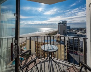 A balcony with a view of the beach and ocean, a round table, and a pool area.