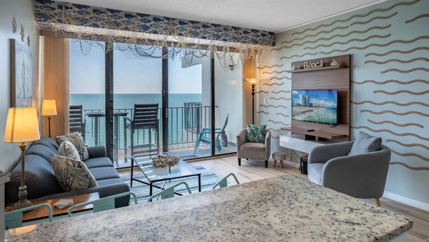 A modern living room with a large TV, seating area, and a balcony with a view of the sea. - Royal Garden Resort 1514 (Murrells Inlet)
