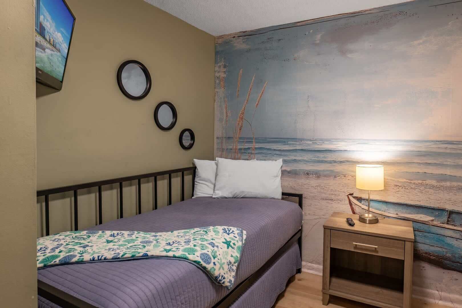 A bedroom with a bed, a nightstand with a lamp, and a wall mural of a beach scene.