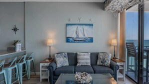 A living room with a sofa, two side tables with lamps, a coffee table with a decorative vase, a painting of a sailboat, and a view of the sea through a balcony. - Royal Garden Resort 1514 (Murrells Inlet)