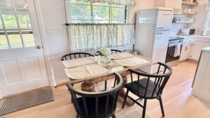 Een eetgedeelte met een houten tafel en stoelen, een raam met gordijnen, en een keuken met witte apparaten.