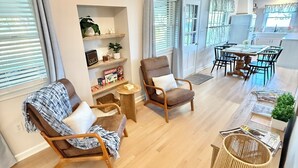 Een moderne woonkamer met een houten vloer, een ronde salontafel, twee fauteuils, een boekenkast, en een eethoek met een tafel en stoelen.