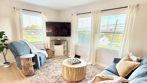Een woonkamer met een blauwe bank, een ronde houten salontafel en een aan de muur bevestigde televisie.