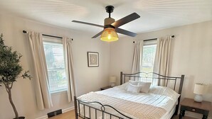 Een slaapkamer met een bed, een plafondventilator, een potplant en een raam met gordijnen.