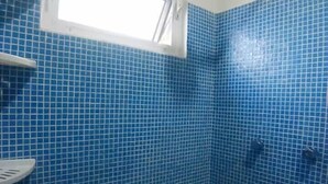 A bathroom with blue tiled walls, a window, a shower, and a white shelf. - En la 96 (Cancún)