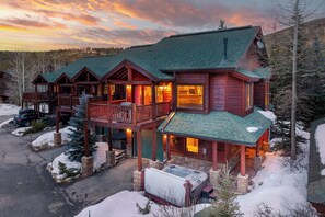 A log cabin with a hot tub, a covered deck, and a view of the surrounding forest.
