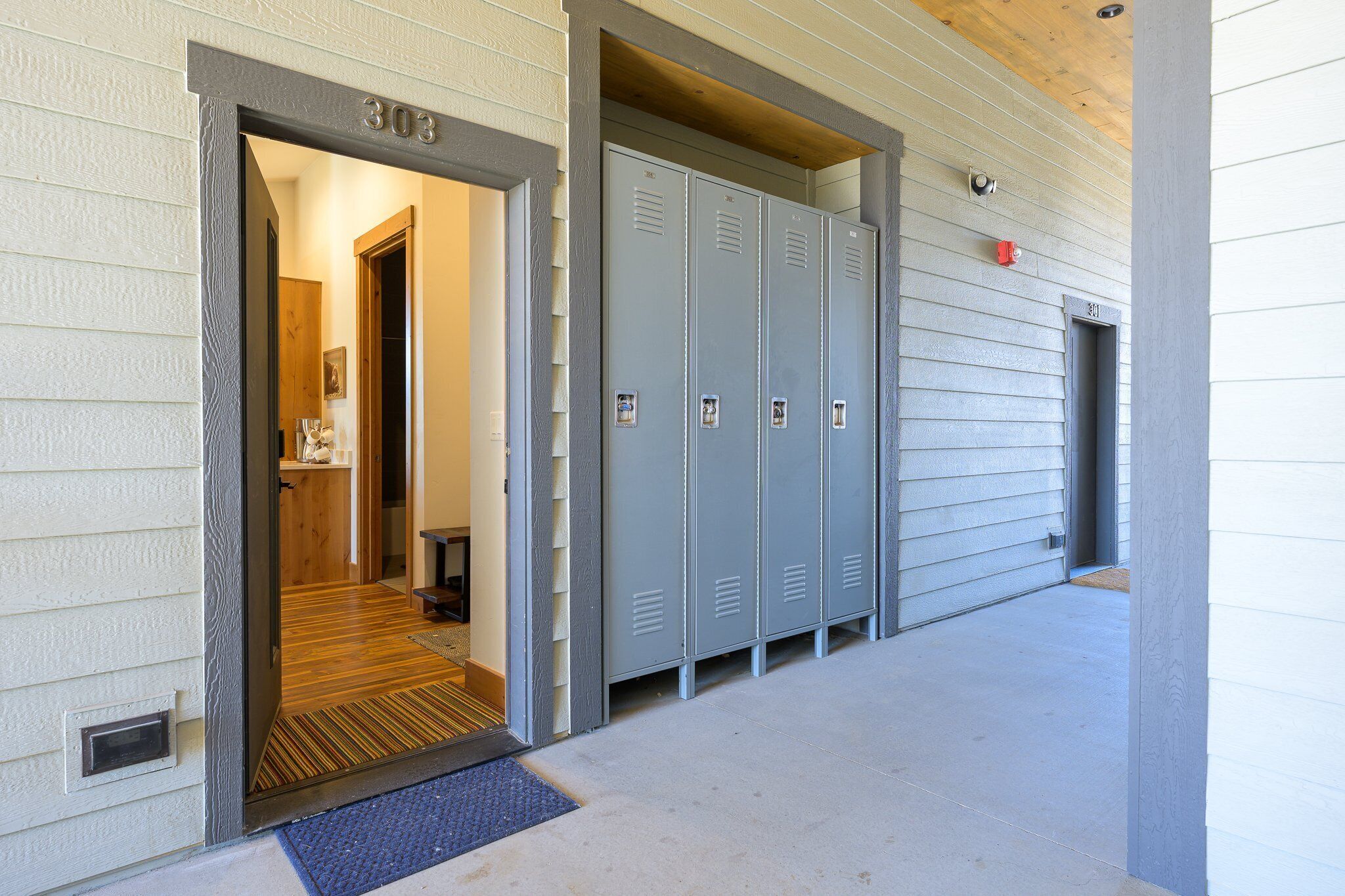 A building entrance with a number 303 sign, a set of grey lockers, and a wooden door leading inside.