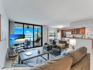 A modern living room with a large TV, comfortable seating, a kitchen area, and a view of a beach. - Waterfront Sunrise View! Marco Island Condo (Marco Island)