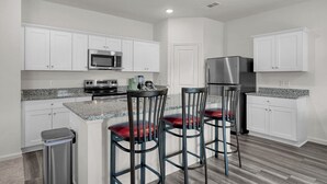 A modern kitchen with white cabinets, granite countertops, stainless steel appliances, and a central island with bar stools. - Beautiful New Build + Central Location + King Bed & Tv's in Every Room (Montgomery)