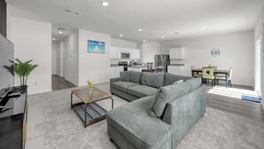 A modern living room with a gray sectional sofa, a coffee table, and a dining area with a table and chairs. - Beautiful New Build + Central Location + King Bed & Tv's in Every Room (Montgomery)