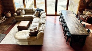 Un salon spacieux avec un grand canapé d’angle, une table à manger en bois et plusieurs chaises. De grandes fenêtres offrent une vue sur l’extérieur.
