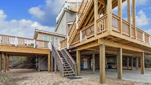 Une propriété située en bord de mer, dotée d’une terrasse en bois, d’un escalier menant à un deuxième étage et d’un sol sablonneux.